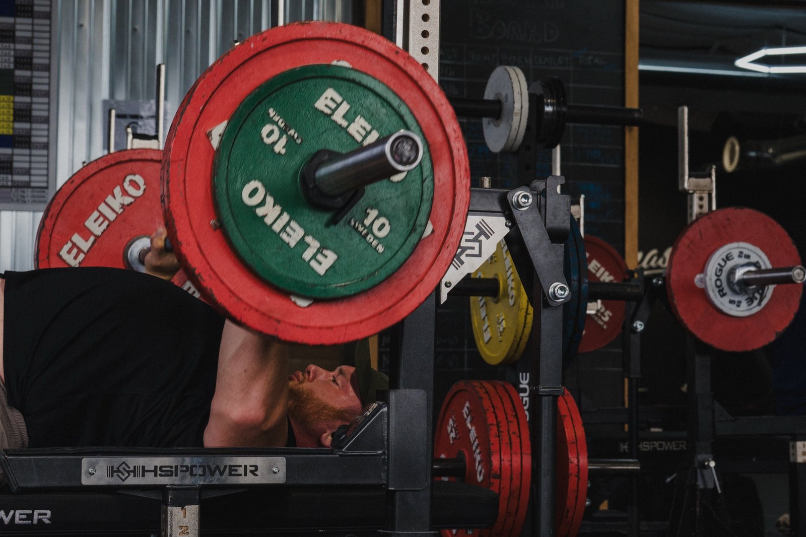 Low-angle photo of a lifter bench pressing a barbell loaded with red and green Eleiko competition plates on the Wolf Dynasty Fitness floor.