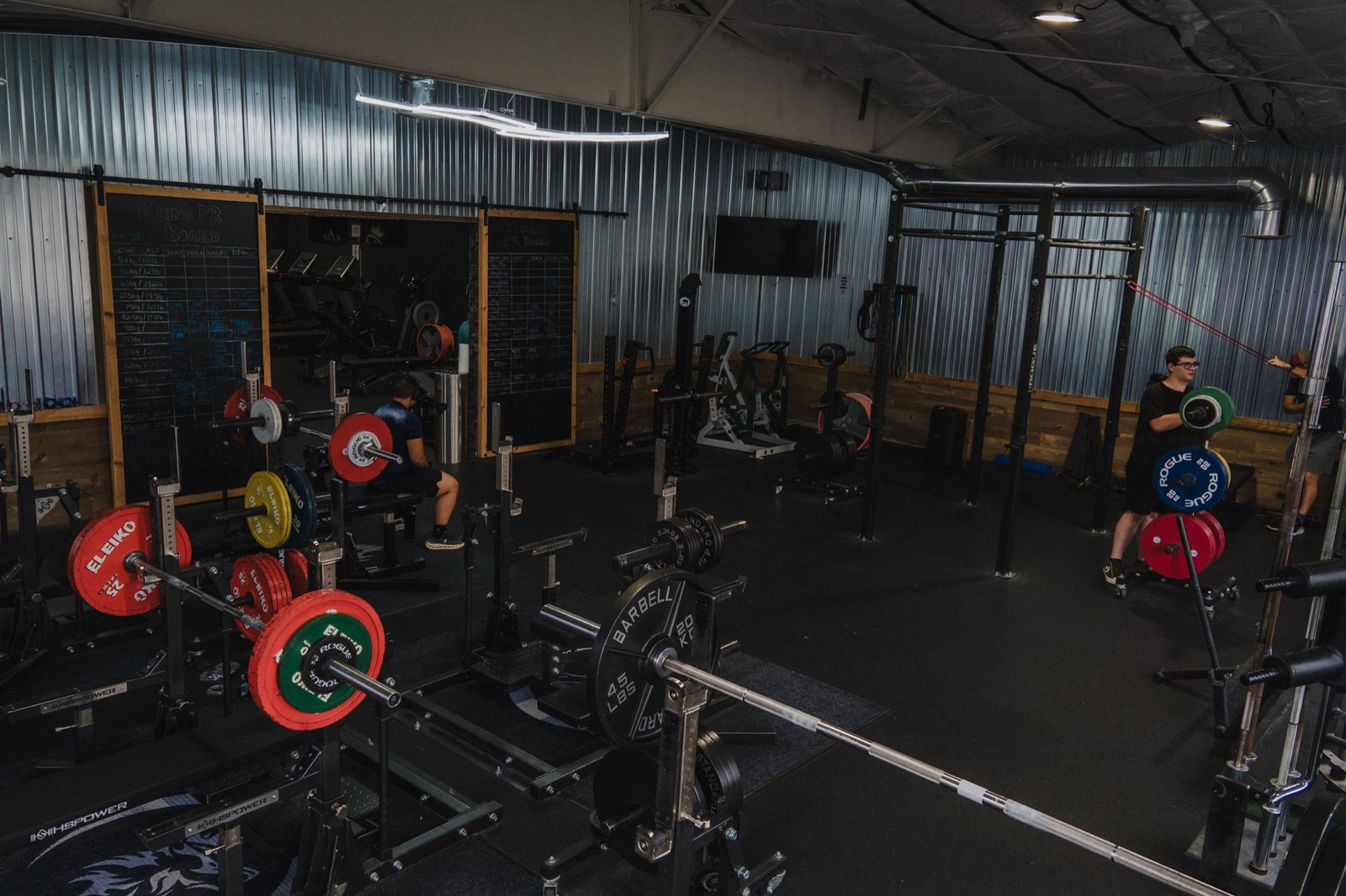 Wide view of the Wolf Dynasty Fitness lifting floor: hand-lettered Men's and Women's PR Board chalkboards on the left, Eleiko and Rogue competition plates racked on multiple stations, members training under warm sodium-toned lighting.