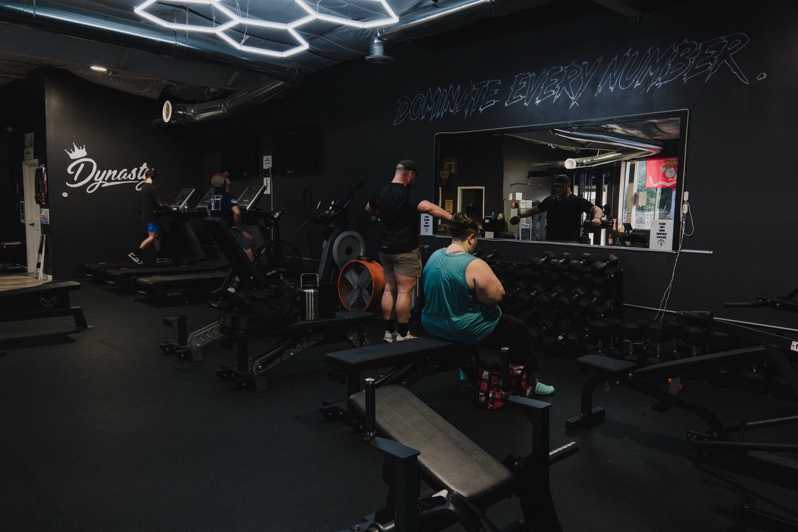 Interior of The Den at Wolf Dynasty Fitness: hand-painted wolf head, crown and crescent moon mural with a hand-lettered script "Dynasty" wordmark on a black wall, a hexagon LED light fixture, "DOMINATE EVERY NUMBER" chalked overhead, and members on the treadmills and cable station.