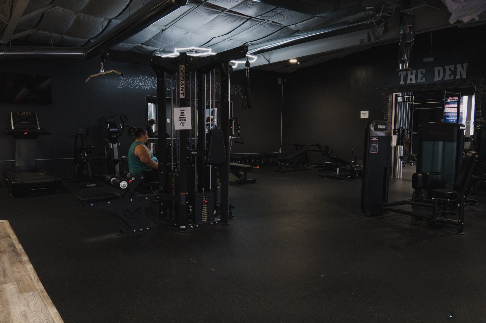A member works through a set on the cable machine in The Den, with "THE DEN" lettering visible through the doorway beyond.