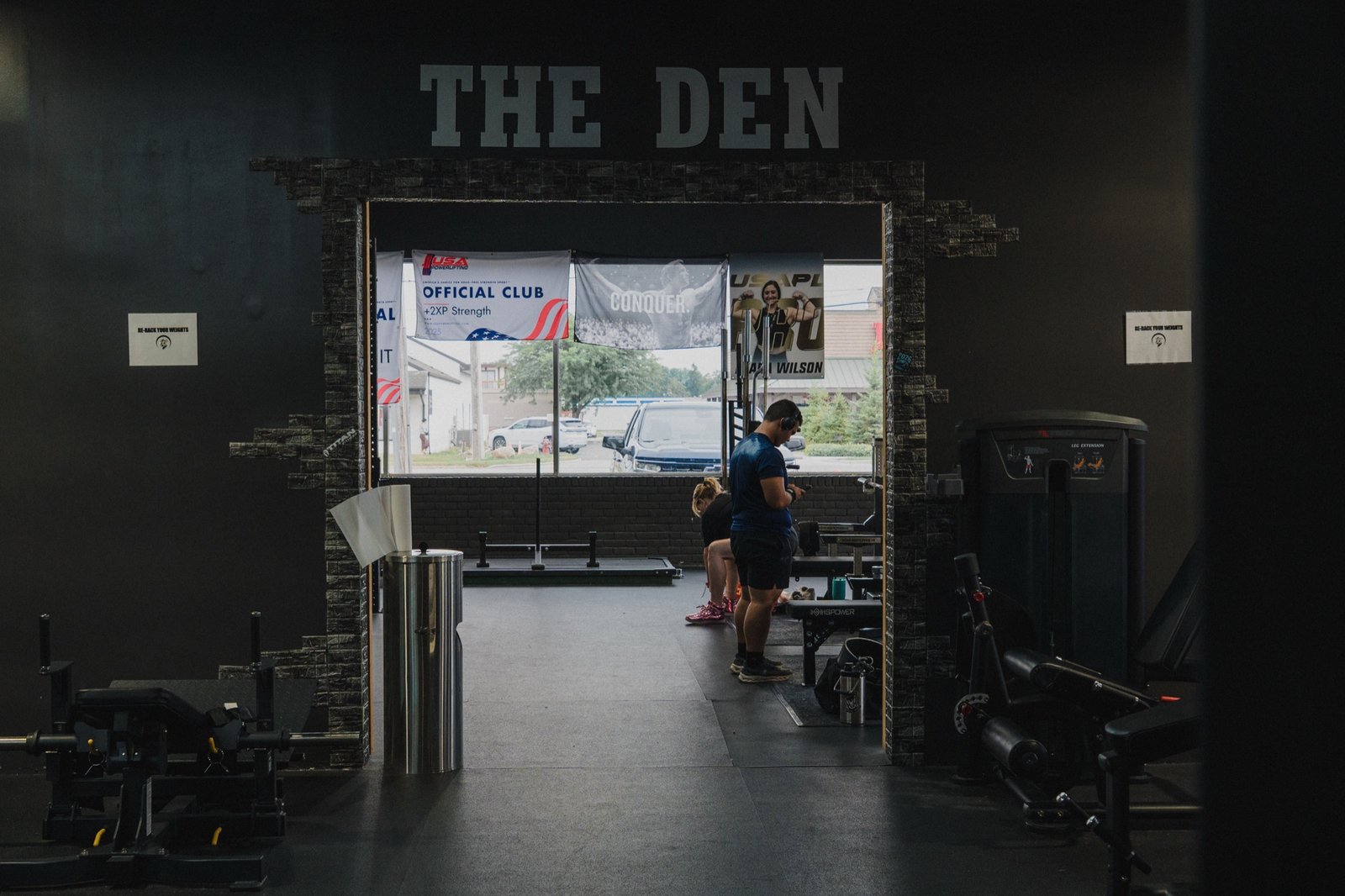 Cinematic doorway shot of "THE DEN" signage above a stone-clad archway at Wolf Dynasty Fitness, USA Powerlifting banners backlit by daylight beyond, two members walking through mid-frame.